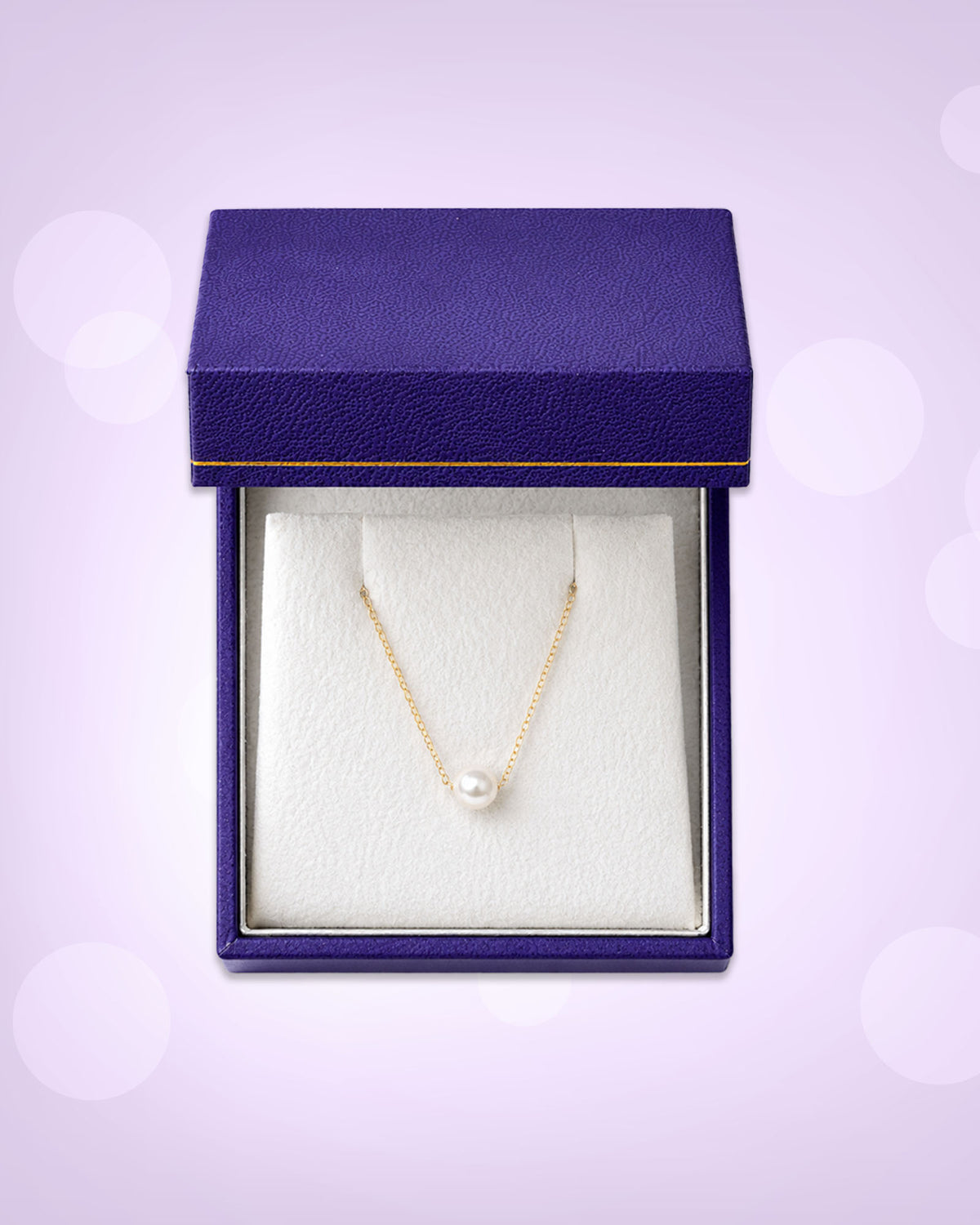 A gold chain necklace with a single white pearl pendant, displayed inside an open textured purple jewelry box with a white insert, set against a light lavender background with soft bokeh circles.