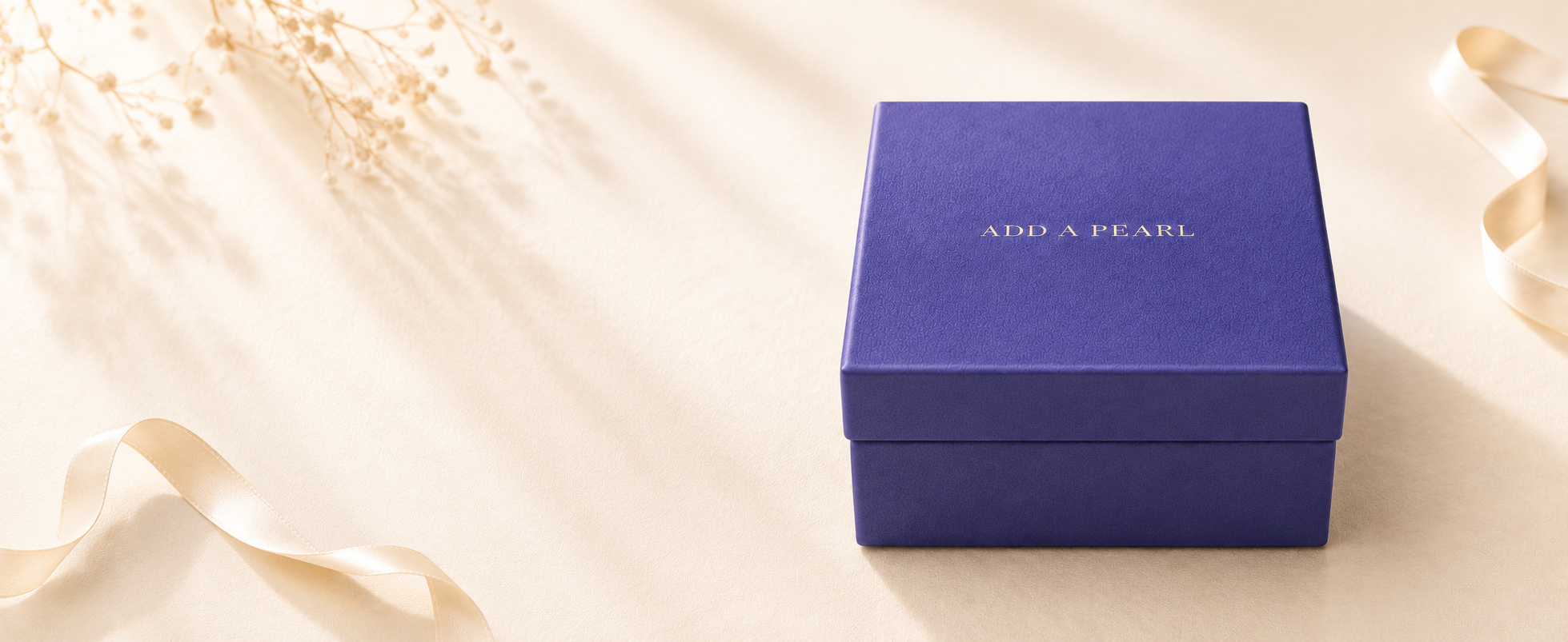 A square royal blue gift box with the words 'ADD A PEARL' printed in white on the lid, styled on a warm cream surface with soft ribbon and dried floral accents.