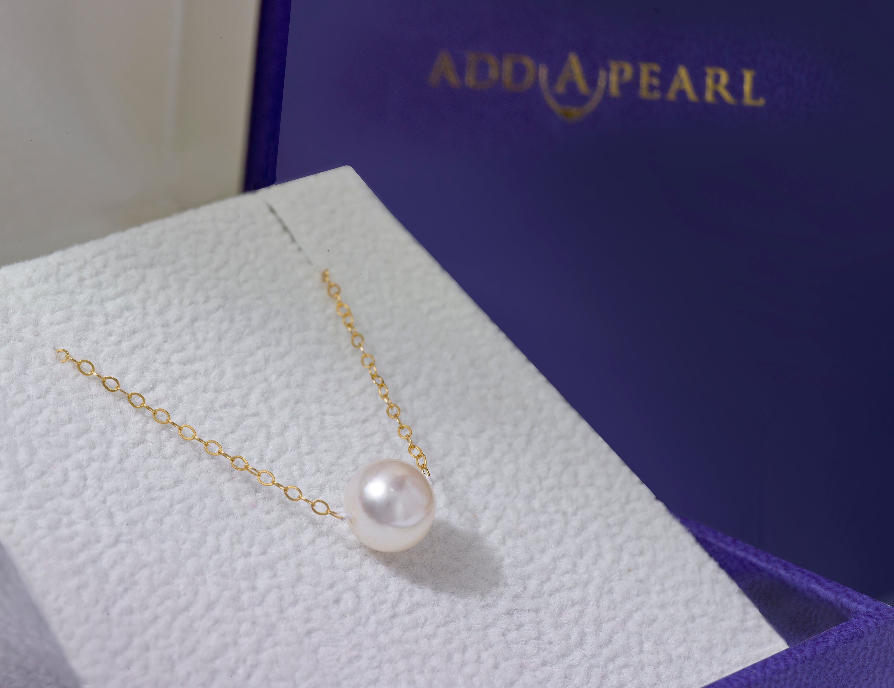 A single cultured pearl on a fine gold chain rests on the white silk cushion of the signature purple Add-A-Pearl gift box of today