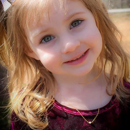 Young girl with blonde hair and blue eyes smiling outdoors wearing a single-pearl gold-chain necklace