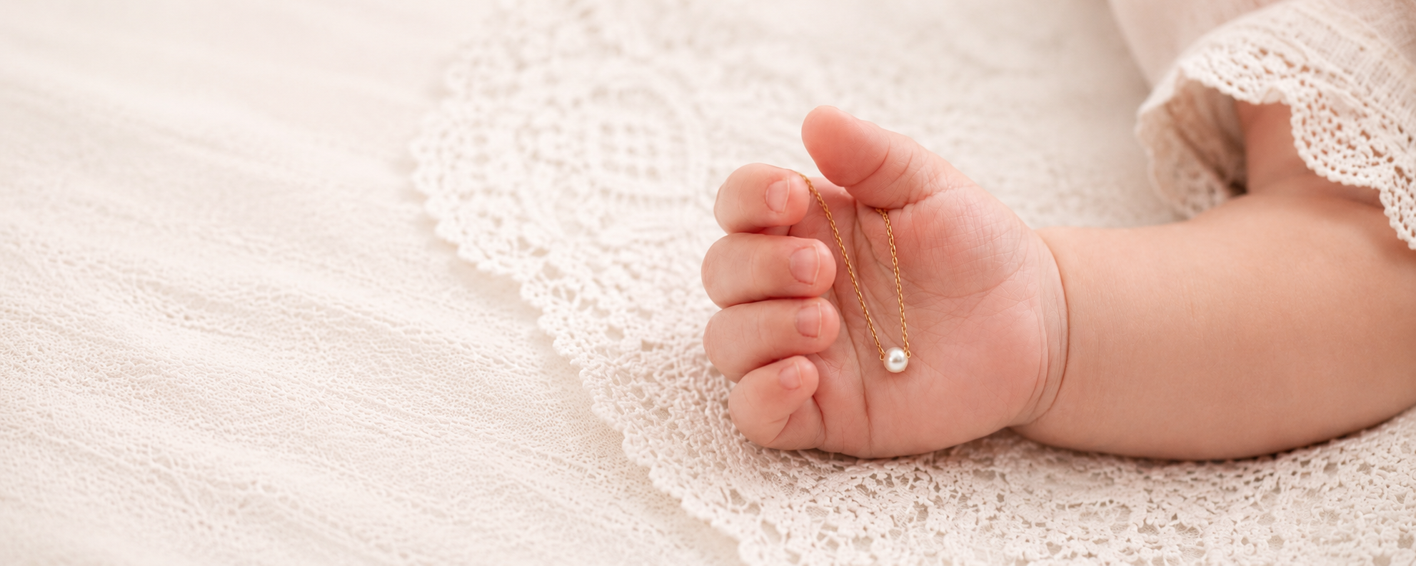 Newborn hand resting on lace with a single pearl on a delicate gold chain
