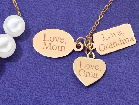 Three engraved gold charms — Love Mom oval, Love Grandma rectangle, and Love Gma heart — on a delicate gold chain with two pearls, against signature Add-A-Pearl purple velvet