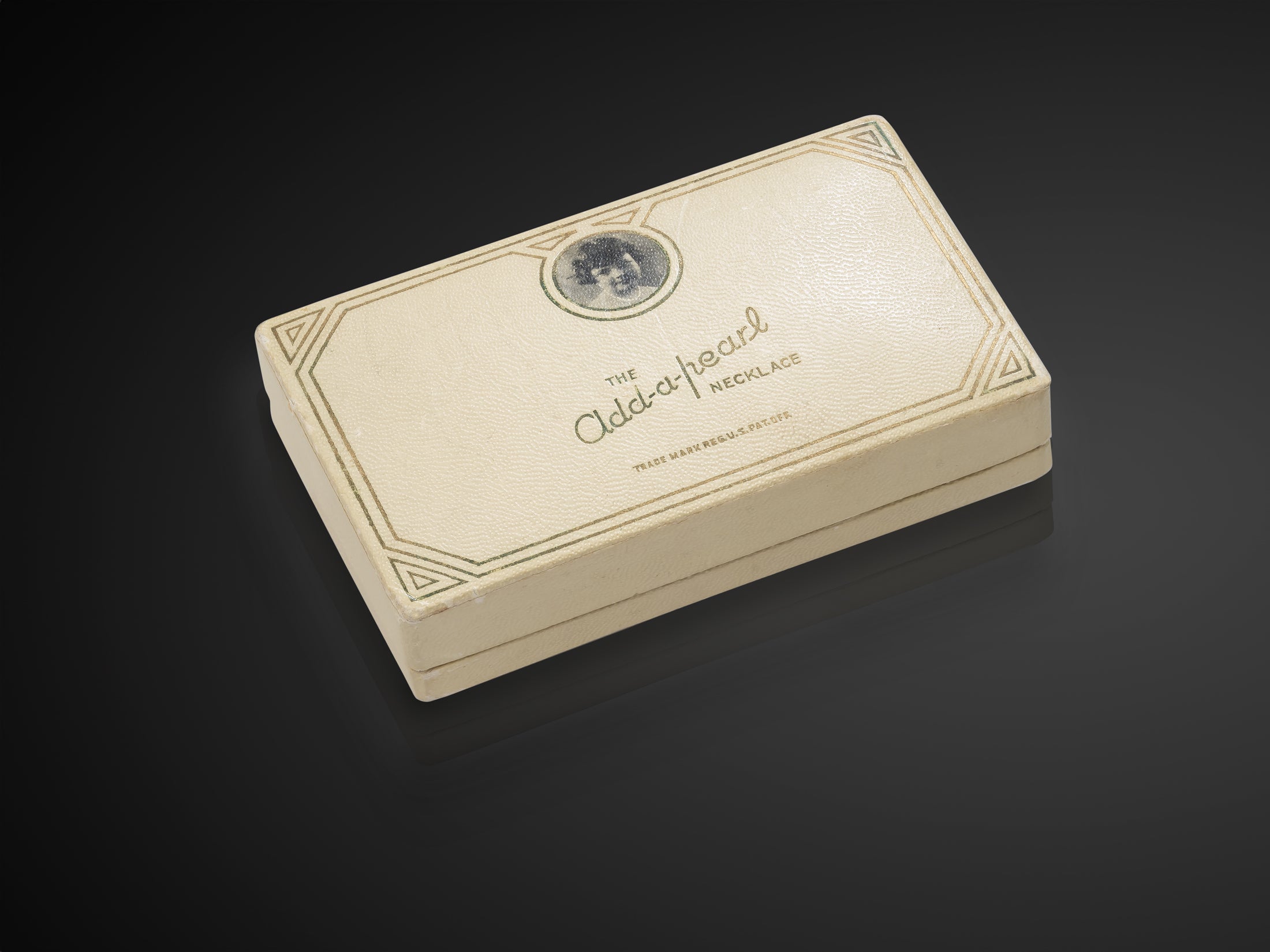 An original Add-A-Pearl gift box from the early twentieth century, photographed against a dark gradient — cream paperboard with gold-and-green Art Deco borders, an embossed portrait medallion, and the cursive Add-a-pearl mark
