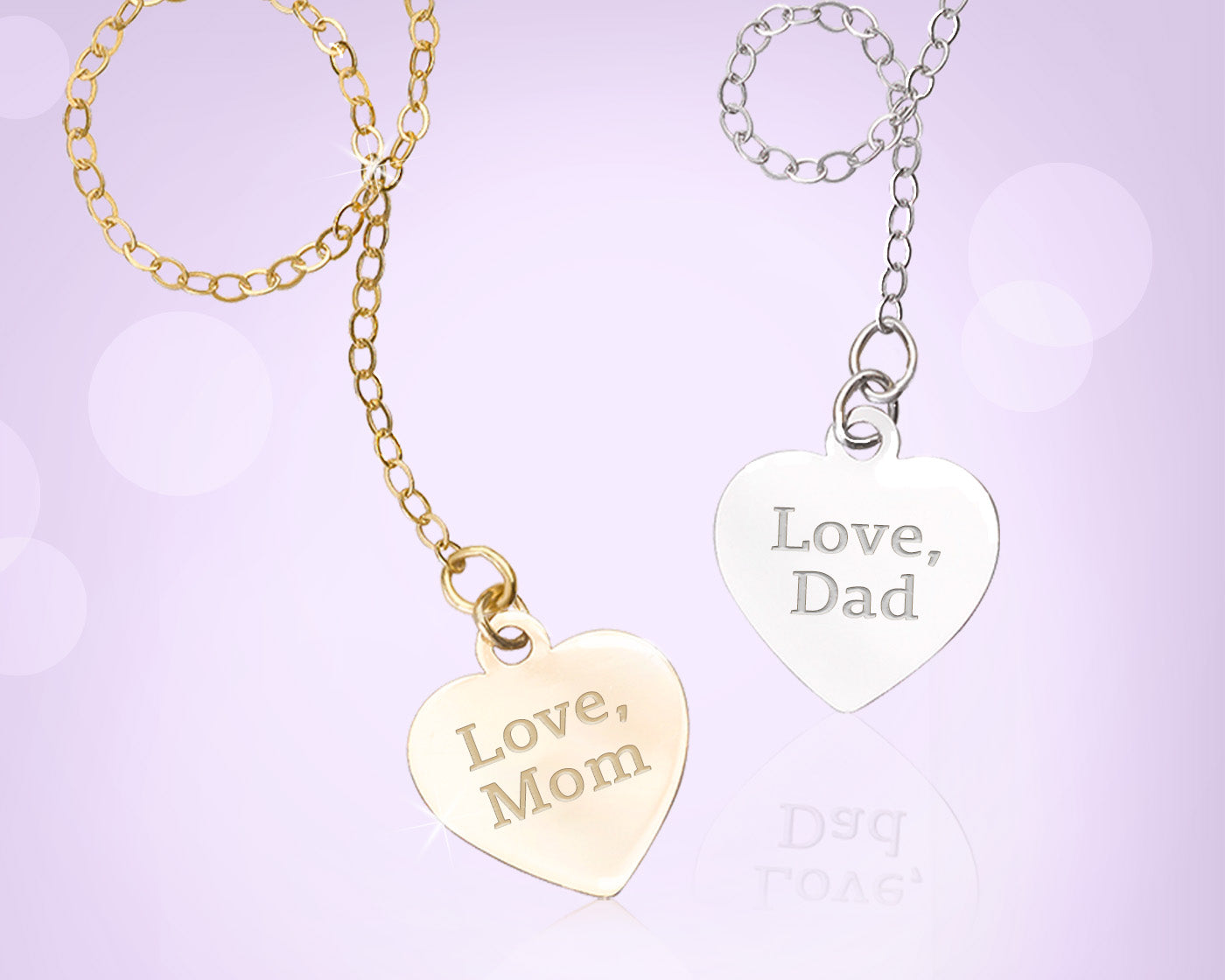 Two heart-shaped pendant necklaces on chains, one gold-toned engraved with 'Love, Mom' and one silver-toned engraved with 'Love, Dad', shown on a soft lavender background.