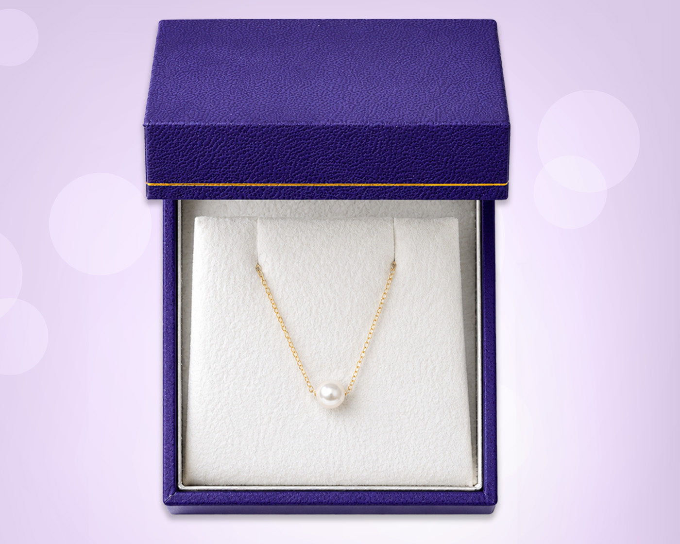 A gold chain necklace with a single white pearl pendant, displayed inside an open textured dark purple jewelry box with a white insert, set against a light lavender background with soft bokeh circles.