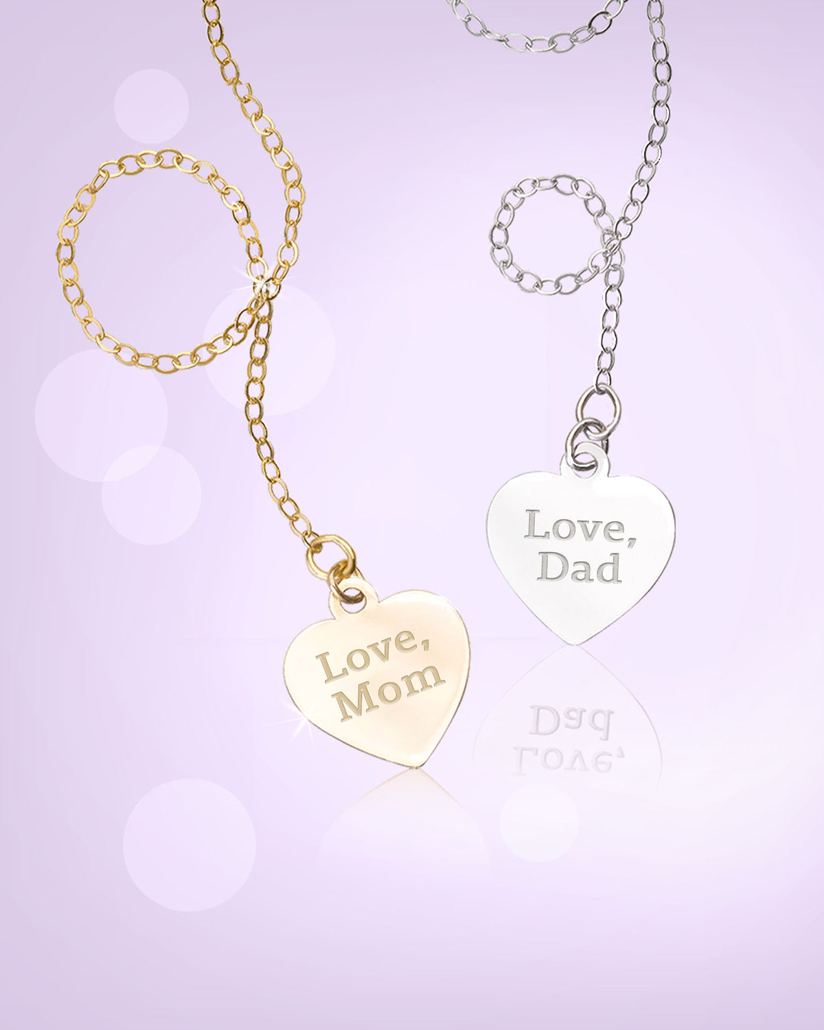 Two heart-shaped pendant necklaces on chains, one gold-toned engraved with 'Love, Mom' and one silver-toned engraved with 'Love, Dad', displayed on a soft lavender background with a faint reflection.
