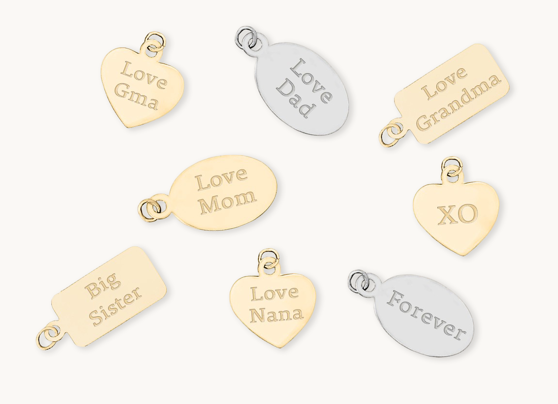 Seven personalized jewelry charms in gold and silver tones, each engraved with a different message such as 'Love Gma,' 'Love Dad,' 'Love Mom,' 'Love Grandma,' 'Big Sister,' 'Love Nana,' 'Forever,' and 'XO,' arranged on a light cream background.