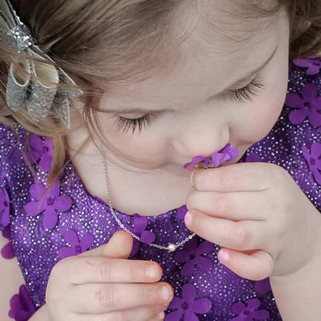 Child in a purple dress wearing a pearl necklace smelling a small flower