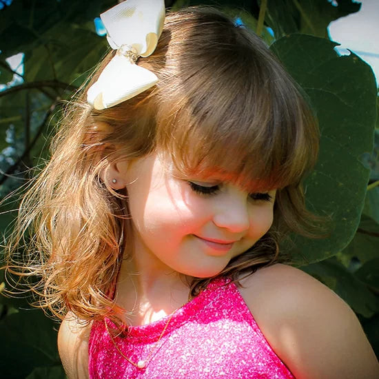Young girl with a large bow in her hair, wearing a pearl necklace, surrounded by green leaves.