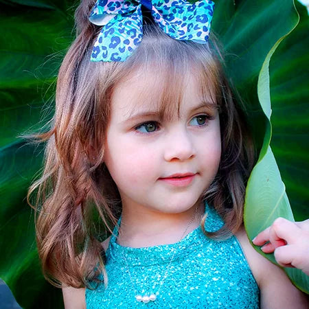 Young girl with a blue bow in her hair, wearing a turquoise dress, standing among green leaves, wearing a graduated pearl necklace
